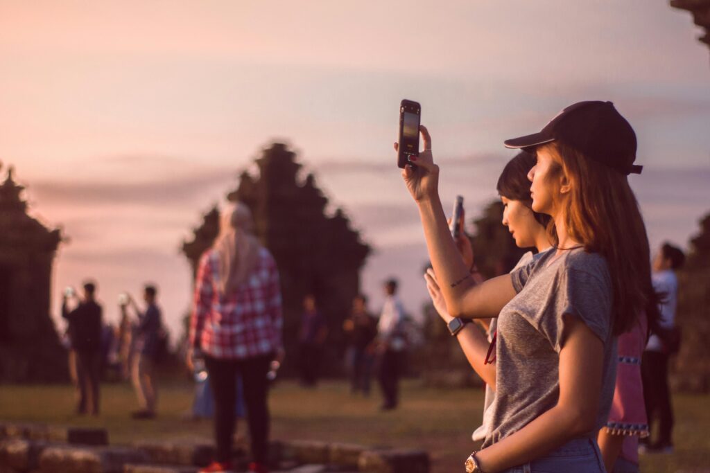 Two young women take photos of a scenic sunset, surrounded by others enjoying the view.